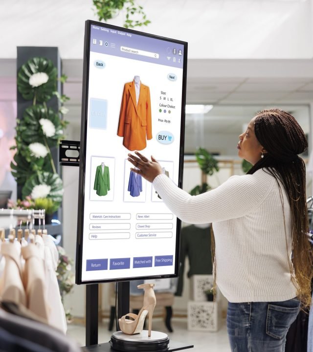 Woman buyer selecting clothes on board, using interactive touch screen monitor in clothing store. Young boutique customer buying fashion items from self ordering kiosk service, retail shop.